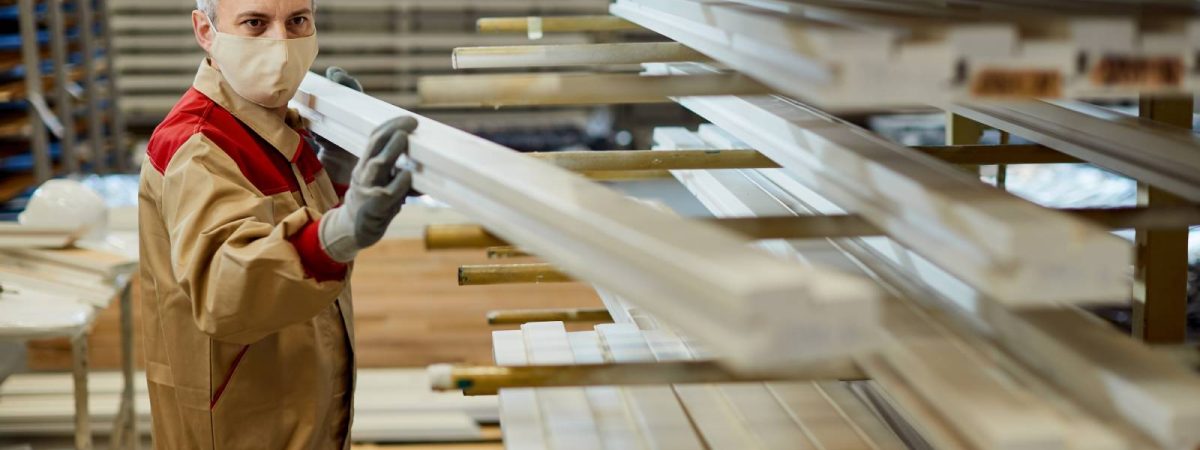 Carpenter taking wood plank from a rack ad wearing face mask while working at workshop during coronavirus pandemic.